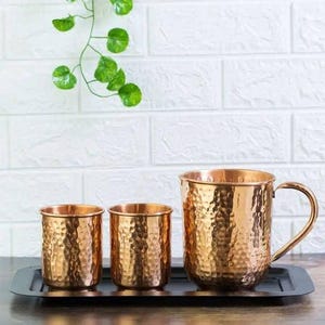 May include: Three copper-coloured hammered metal mugs and cups of varying sizes sit on a black tray. The largest mug has a handle. The background features a white brick wall and a green vine.