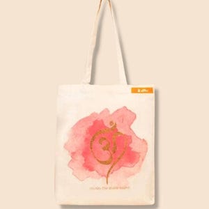 Puede incluir: Bolso tote de color crema con un diseño de acuarela rosa y un símbolo Om dorado. El bolso tiene asas largas y el texto "(AUM) The divine sound". Hecho de tela clara.