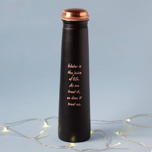 May include: A black water bottle with a copper-coloured lid. The bottle has a slender design and features the text "Water is the juice of life. As we treat it, so does it treat us." in a gold colour.