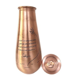 May include: A copper-toned water bottle with a matching lid. The bottle has a tapered shape and features etched text and a floral design. The lid is round with a raised emblem. The text on the bottle reads, "You hold the water you drink..." and the word "Sadhguru".
