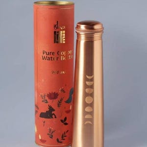 Puede incluir: Una botella de agua de cobre con un diseño de fases lunares y un contenedor cilíndrico a juego. La botella está etiquetada como "Pure Copper Water Bottle" y tiene una capacidad de 710 ml. El contenedor es naranja con detalles dorados e ilustraciones.
