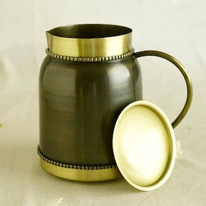 May include: A vintage-style metal jug with a handle and a lid. The jug has a dark bronze body with gold-coloured accents around the top, base, and handle. The lid is also gold-coloured. The jug is designed to hold liquids.