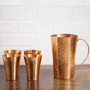 May include: A copper-coloured hammered metal jug with a handle and four matching tumblers. The jug and tumblers have a textured surface and are arranged on a dark wooden surface against a white brick wall.