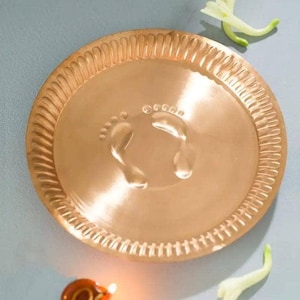 May include: A circular copper-toned plate with a textured edge, featuring a central imprint of two feet. A small, lit oil lamp and white flowers are also present, possibly indicating a cultural or religious context.