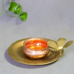 May include: A brass candle holder featuring a small copper-coloured bowl with a lit candle. The candle holder rests on a round brass plate, with a brass flower-shaped accent. The background is a neutral grey.