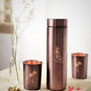 May include: A set of copper-coloured stainless steel water bottle and two tumblers. The bottle has an etched design of a figure and the word "Adiyogi". The tumblers and bottle have a lotus flower design.