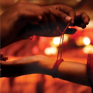 Puede incluir: Primer plano de un hilo rojo atado alrededor de una muñeca. La imagen muestra dos manos, una atando el hilo y la otra sosteniendo la muñeca. El fondo tiene un tono cálido, rojizo-anaranjado.