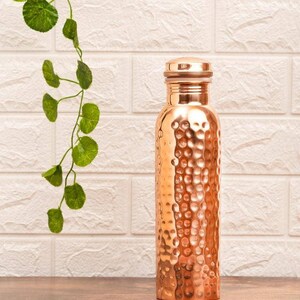 May include: A copper water bottle with a textured, hammered finish. The bottle has a screw-on cap and a cylindrical shape. The background features a white brick wall and a green vine. The bottle is approximately 25 cm tall.