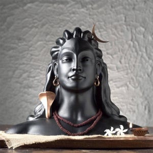 Puede incluir: Busto negro de una deidad con pelo largo, pendientes y una luna creciente. Una figura de serpiente y un collar de cuentas adornan la escultura. Dos flores blancas y una pequeña lámpara de aceite están frente al busto.