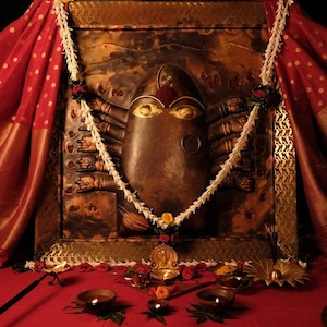 Puede incluir: Un icono religioso de bronce con múltiples brazos y detalles dorados, adornado con una guirnalda y tela roja. Velas encendidas y pequeñas estatuas están dispuestas sobre una superficie roja, creando una atmósfera devocional.