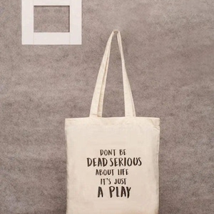 Puede incluir: Bolso tote de lona color crema con el texto negro "DON'T BE DEAD SERIOUS ABOUT LIFE IT'S JUST A PLAY". El bolso tiene asas largas y cuelga contra una pared gris texturizada con un marco cuadrado blanco.