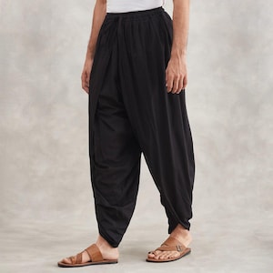 Puede incluir: Pantalones harem negros con un diseño suelto y drapeado. Los pantalones tienen una cintura fruncida y un ajuste relajado, con una pernera ancha y fluida. El modelo lleva sandalias de cuero marrón. El fondo es una pared neutra y texturizada.
