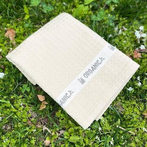 Puede incluir: Toalla plegada de color crema con un tejido texturizado, sobre hierba verde. Una banda blanca con la palabra "ORGANIC" envuelve la toalla. La toalla parece estar hecha de un material suave y natural.