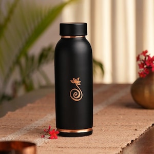 May include: A black water bottle with copper-coloured accents. The bottle has a copper-coloured floral design. The bottle is cylindrical and has a black cap. The bottle is on a brown surface.