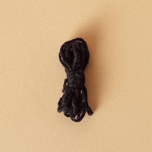 Puede incluir: Un pequeño ovillo de hilo negro, bien enrollado y anudado, se encuentra sobre un fondo beige. El hilo parece estar hecho de un material suave y texturizado, con hebras sueltas visibles en la parte inferior. Adecuado para proyectos de manualidades.
