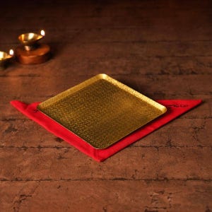 May include: A square, gold-colored tray with a textured surface rests on a red cloth. The tray is centered on a dark brown wooden surface. In the background, a small, lit candle holder is visible.
