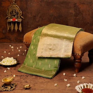 May include: A green and gold sari draped over a small stool. The sari has a gold border and gold floral patterns. A small brass lamp and a bowl of flowers are in the foreground. The background is a dark brown.