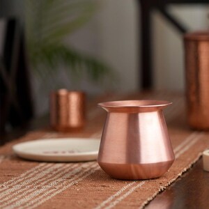 May include: A copper-coloured metal pot with a flared rim and a rounded body. The pot sits on a brown woven surface, with a small white plate and other copper items in the background. The pot has a shiny, reflective surface.