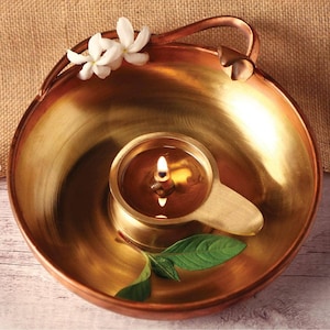 May include: A brass oil lamp with a lit wick sits inside a larger brass bowl. The bowl has a handle and is decorated with white flowers and a green leaf. The lamp is filled with oil, and the flame is bright.