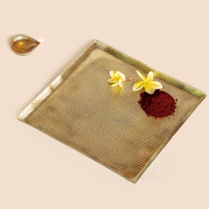Puede incluir: Una bandeja cuadrada dorada con una superficie texturizada. Dos flores amarillas y un montón de polvo rojo están en la bandeja. Un pequeño plato en forma de lágrima con líquido está en la esquina superior izquierda.