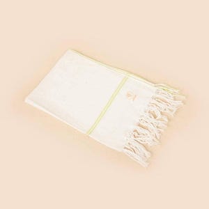 Puede incluir: Toalla rectangular blanca con una raya verde claro y flecos. La toalla tiene un pequeño diseño bordado en la esquina. La toalla está doblada y descansa sobre un fondo melocotón claro.