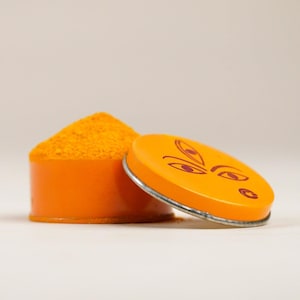 Op de afbeelding: Een oranje container met een deksel met een gestileerd gezichtsontwerp. De container bevat een hoop felgeel poeder. Het deksel staat iets open en onthult de inhoud. De container staat op een wit oppervlak.