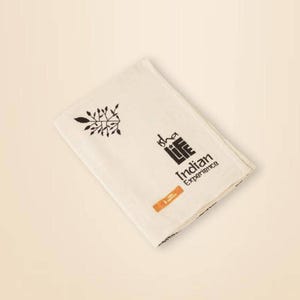 Puede incluir: Paño plegado de color crema con diseños estampados en negro y naranja. El paño presenta una ilustración en forma de rama, el texto "Isha Life Indian Experience" y un pequeño rectángulo naranja.