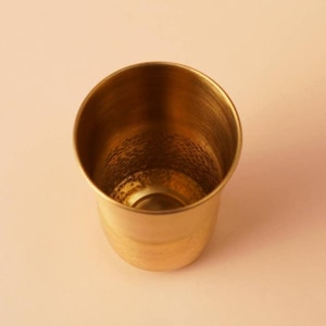 May include: A small, gold-coloured metal cup with a hammered texture on the inside. The cup has a rounded base and a slightly flared rim. The cup is empty and sits on a light pink surface.