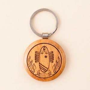 Puede incluir: Un llavero redondo de madera con un aro plateado. El colgante de madera presenta un diseño grabado de una figura estilizada con llamas a ambos lados. El diseño es de color marrón oscuro sobre madera marrón claro.