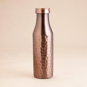 May include: A copper-coloured water bottle with a hammered texture. The bottle has a rounded body and a narrow neck with a matching copper cap. The bottle is made of metal and has a shiny finish.