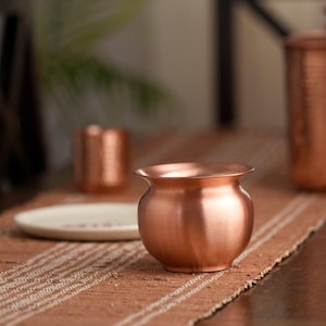 May include: A small, copper-coloured pot with a rounded body and flared rim. The pot sits on a brown and white striped runner, with a blurred background featuring other copper items and a white plate.