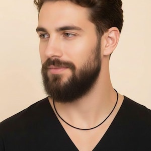 Op de afbeelding: Een eenvoudige zwarte ketting wordt gedragen. De ketting is een dun koord. De persoon op de foto draagt een zwarte top. De achtergrond is effen lichtbeige.