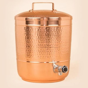 Puede incluir: Un dispensador de agua de cobre con textura martillada. El dispensador tiene una tapa con asa y un grifo cerca de la base. El acabado de cobre le da un aspecto cálido y rústico. Está diseñado para contener y dispensar agua.