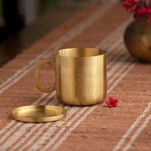 May include: A gold-coloured metal mug with a matching saucer. The mug has a handle and an etched sun and moon design. The mug and saucer sit on a brown and white striped surface. A small red flower is next to the mug.