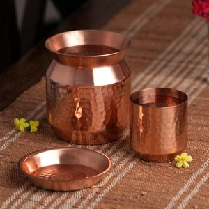 May include: A set of three copper home decor items: a hammered-finish vase, a tumbler, and a small dish. The copper pieces have a warm, reddish-brown hue and are arranged on a textured, neutral-toned surface.
