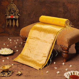 May include: A golden yellow silk sari is draped over a small, brown upholstered stool. The sari is folded and rolled, with tassels visible. The scene is decorated with small white flowers and a brass lamp.