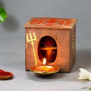 Puede incluir: Un objeto decorativo en forma de caja de color cobre con una abertura ovalada que revela la cara de una deidad. Un tridente dorado y una pequeña lámpara de aceite con una llama encendida están en la parte delantera. La parte superior de la caja tiene marcas rojas.