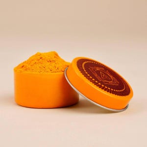 Puede incluir: Un recipiente naranja abierto lleno de polvo amarillo brillante, junto a una tapa naranja con un diseño geométrico marrón. La tapa tiene un borde plateado. El fondo es de color beige claro liso.