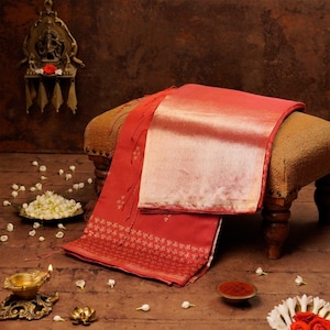 May include: A red and gold sari is draped over a small stool. The sari has a gold border and intricate gold patterns. The background includes a small decorative shelf, a bowl of flowers, and a small oil lamp.