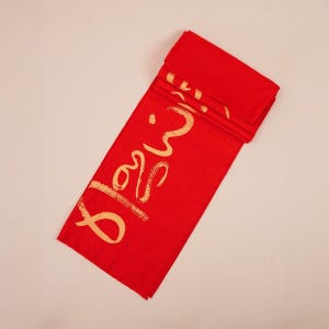 Puede incluir: Un camino de mesa de tela roja doblada con texto dorado. El texto dice "Bless". El camino de mesa es rectangular y parece estar hecho de un material liso. El fondo es de un color sólido y neutro.