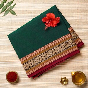 May include: A folded dark green textile with a decorative border featuring a repeating pattern in gold, black, and red. A red flower rests on the fabric. A small bowl of red powder and a brass oil lamp are also present.