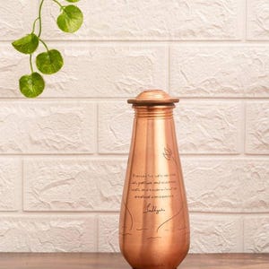 May include: A copper water bottle with a wooden lid. The bottle has etched floral designs and text that reads "Possess the wisdom with patience and resilience..." The bottle is approximately 25 cm tall.