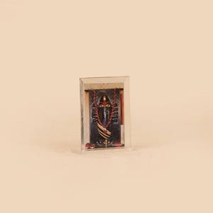Puede incluir: Icono religioso enmarcado que presenta una deidad oscura con detalles rojos y un collar de cuentas. El icono se muestra en un soporte rectangular transparente, sobre un fondo neutro.