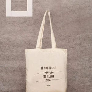 Puede incluir: Bolso tote color crema con asas largas, con la cita "If you resist change you resist life" en escritura negra. Un marco rectangular blanco está en la esquina superior izquierda. El bolso está hecho de tela resistente.