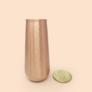 May include: A copper-coloured, hammered metal vase with a matching round lid. The vase has a tall, slender shape and a textured surface. The lid is a flat, circular piece of brass. The background is a solid, light peach colour.