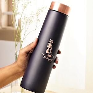May include: A dark blue insulated water bottle with a copper-coloured lid. The bottle features a rose gold graphic of a spiritual figure with the word "Adiyogi" below. The bottle is held by a hand.