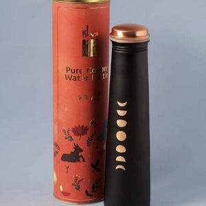 May include: Black water bottle with a copper lid and a series of moon phase designs. The bottle is next to a cylindrical orange container with the text "Pure Copper Water Bottle" and floral and animal illustrations.