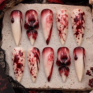 May include: A set of ten stiletto-shaped press-on nails featuring an off-white base with various red blood-splatter patterns, drips, and three-dimensional sculpted floral accents in deep red, arranged in two rows on a textured stone surface.