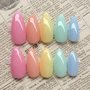 May include: A set of ten almond-shaped press-on nails arranged in two rows of five. The colors include glossy pink, peach, pale yellow, mint green, and light blue. Each nail features a smooth, solid finish with a reflective surface and rounded tips.