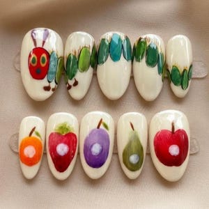May include: A set of ten artificial nails with a cream-colored base. The top row features a caterpillar design and green leaves. The bottom row displays orange, red, purple, green, and red fruit designs. Each nail is oval-shaped.